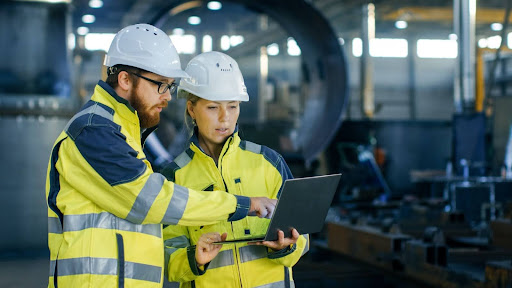 Two people outfitted in protective equipment looking at a laptop in a manufacturing plant.